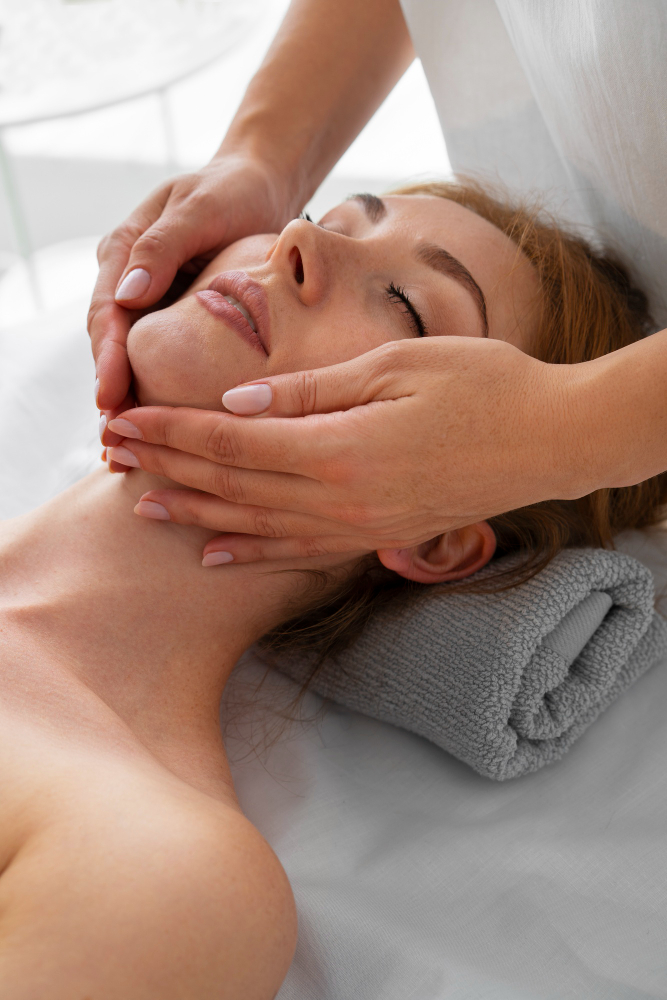 En kvinna som ligger på en grå handduk och blir masserad i ansiktet av två händer.